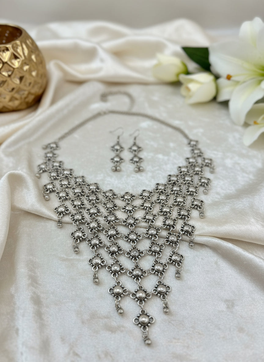 Silver statement necklace with matching earrings, featuring intricate floral designs and a layered look.