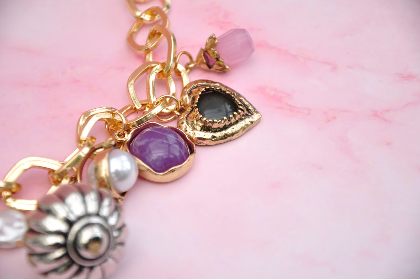 Gold charm bracelet with purple and black gemstones and pearl accents on a pink marble background.