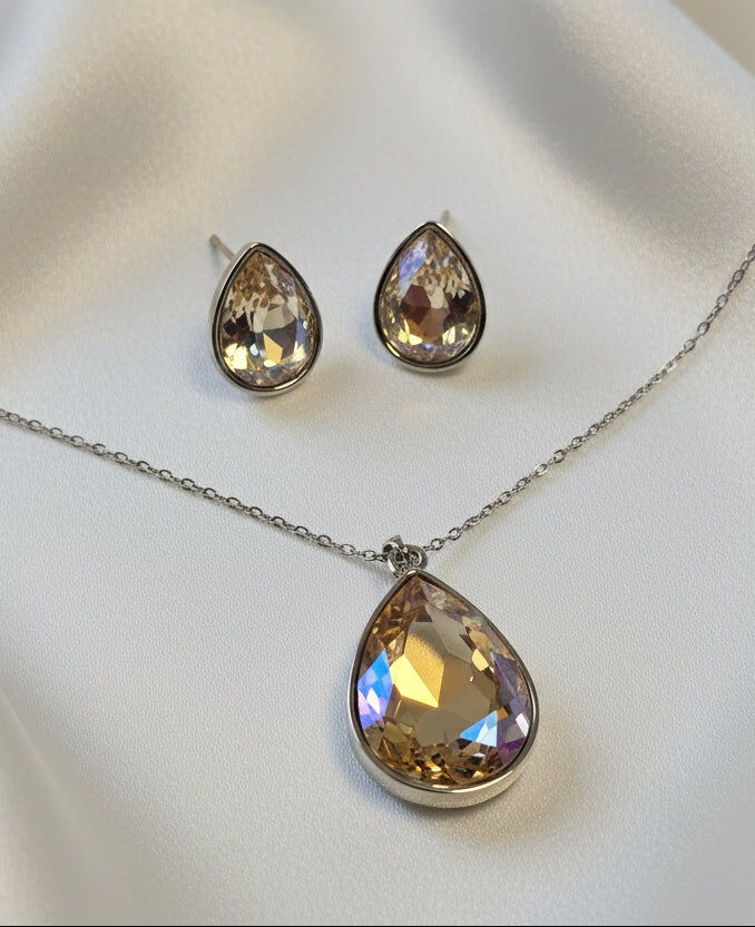 Elegant gold-tone jewelry set with teardrop cut crystal pendant and matching earrings on satin background.