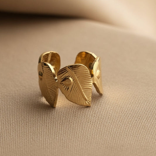 A gold-toned open cuff ring with three leaf-shaped elements, photographed on a soft beige textured surface.