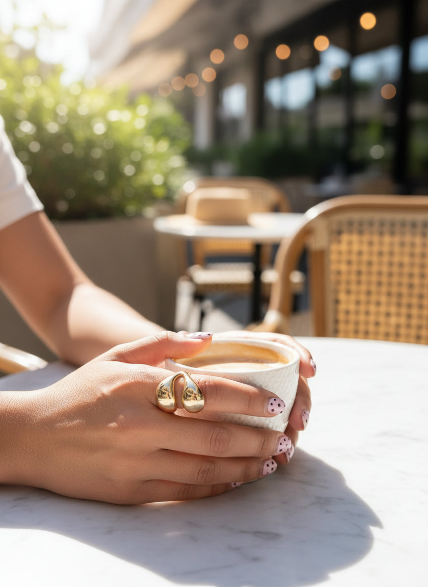 Woman holding a white coffee cup, wearing a stylish gold ring, with a sunny café backdrop.