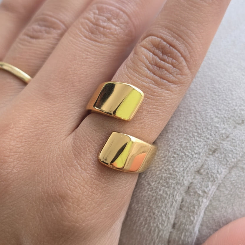 A close-up of a hand wearing two chunky, polished gold rings with a square, beveled edge design.