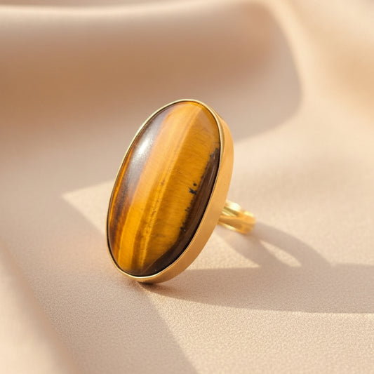 A gold ring featuring a large oval tiger's eye gemstone with bold brown and yellow tiger's eye stripes, resting on a reflective dark surface.